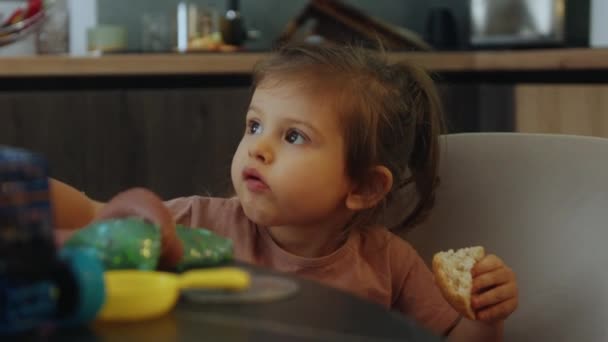 Portré gyönyörű lány, hogy eszik kenyeret játék közben a konyhaasztalnál. - Felvétel, videó
