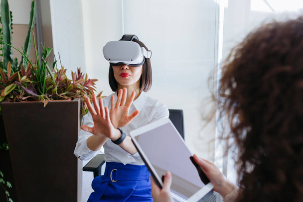 siedzi Azji kobieta w białej koszuli mając na sobie widz zwiedzanie wirtualnej rzeczywistości i jej kolega pracuje z tabletem - Zdjęcie, obraz