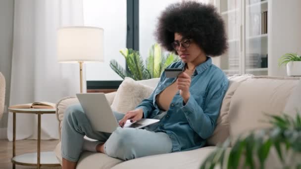 afro-américaine ethnique fille faire paiement en ligne tenant carte de crédit bancaire en utilisant un ordinateur portable moderne à la maison assis sur canapé maison femme femme client acheteur ordinateur shopping internet bancaire - Séquence, vidéo