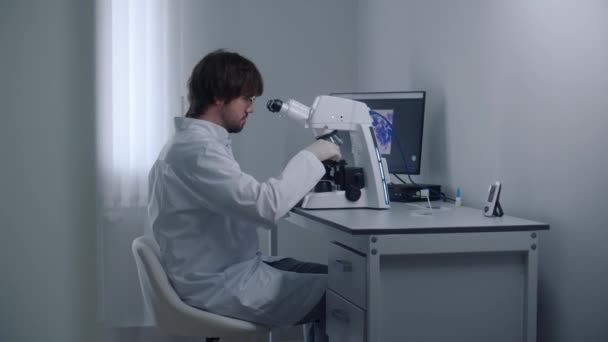 Чоловічий вчений дивиться під мікроскопом, вивчає і аналізує бактерії або зразки крові в сучасній лабораторії. Професійний хімік працює в передовій лабораторії, займається біотехнологією або фармацевтичними дослідженнями. - Кадри, відео