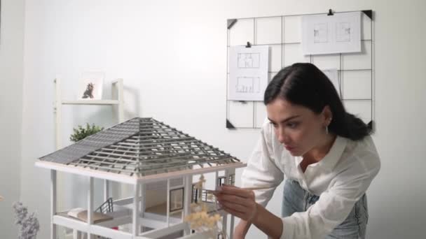 Detailní záběr mladé krásné inteligentní inženýrky se zaměřuje na měření modelu domu pomocí tužky v moderní kanceláři s nákresem svlečeným na stěně. Tvůrčí koncept bydlení a designu. Neposkvrněné. - Záběry, video