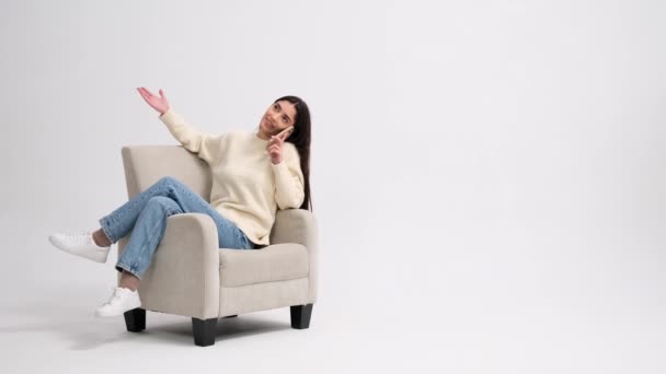 Femme gaie parle au téléphone et se détendre dans un fauteuil sur un fond blanc. Technologie numérique, conversation en ligne, concept d'activité de loisirs. - Séquence, vidéo