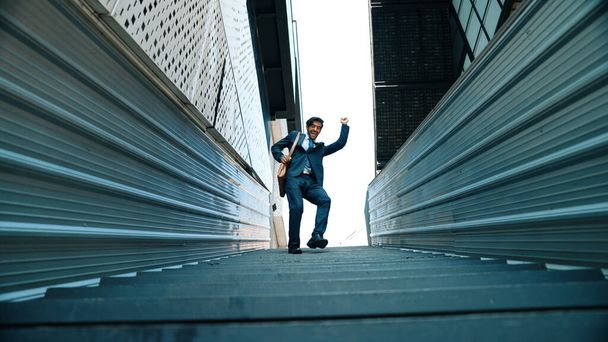Low angle view of young smiling business man dance between building at center point. Skilled executive manager wearing suit and suitcase and moving to music. Happy investor moving to music. Exultant. - Photo, Image
