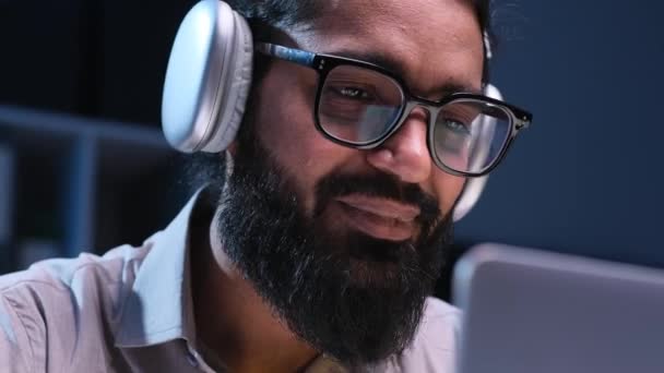 Sikeres indiai üzletember fülhallgatót visel zenét hallgat és énekel, miközben laptoppal dolgozik késő esti irodájában. Vállalkozó élvezi a zenét. - Felvétel, videó