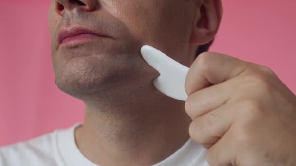 Oříznutý záběr obličeje muže středního věku masírujícího bradu a tváře s nástrojem gua sha izolovaným ve studiu - Záběry, video