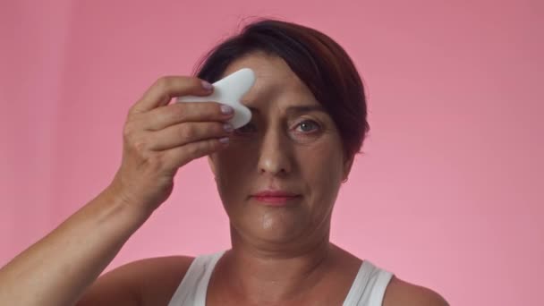 Moyen gros plan de femme blanche mature caucasienne en haut faisant massage facial avec gua sha outil et en regardant la caméra - Séquence, vidéo