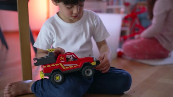 Petit garçon jouant avec camion de pompiers jouet de véhicule sur le sol avec sœur en arrière-plan. Objet de retenue d'enfant focalisé à la main - Séquence, vidéo