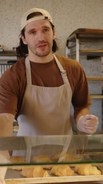 Tiro vertical del trabajador de panadería masculina mostrando surtido de pastelería en pantalla de vidrio y dando consejos al cliente - Metraje, vídeo