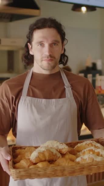 Vertikální portrét mladého pekaře v zástěře držícího tác plný čerstvě upečených croissantů a pózujícího pro kameru s úsměvem - Záběry, video