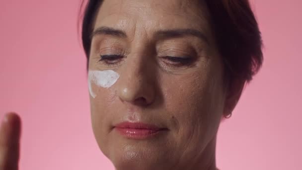 Primer plano de la alegre mujer de mediana edad de etnia caucásica utilizando bajo la crema de levantamiento de ojos para la piel madura en el fondo rosa - Metraje, vídeo
