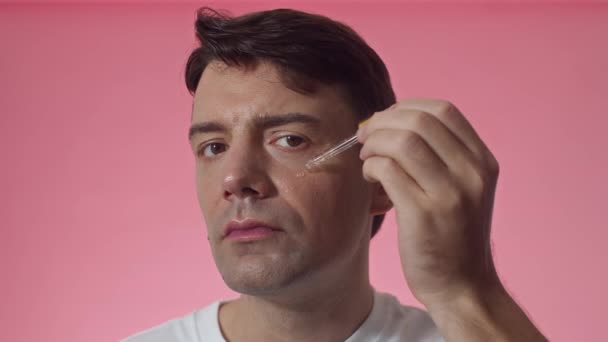 Κοντινό πλάνο ελκυστικού μεσήλικα άνδρα καυκάσιας εθνικότητας που εφαρμόζει ορό ανύψωσης κάτω από μάτια απομονωμένα σε ροζ φόντο - Πλάνα, βίντεο
