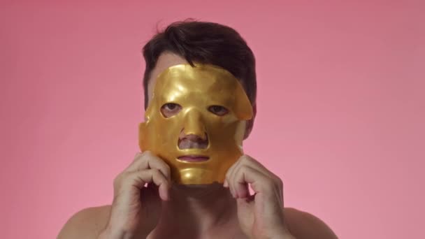 Στιγμιότυπο από ενήλικο άνδρα με καστανά μάτια και σκούρα μαλλιά χρησιμοποιώντας κίτρινη μάσκα φύλλων υδρογέλης στο πρόσωπο που απομονώνεται σε ροζ φόντο - Πλάνα, βίντεο