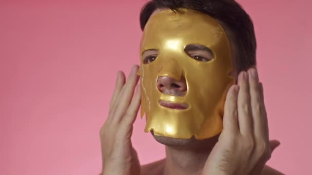 Close up de homem de meia-idade com olhos castanhos colocando máscara de folha de colágeno de cor amarela no rosto em fundo rosa, espaço de cópia - Filmagem, Vídeo