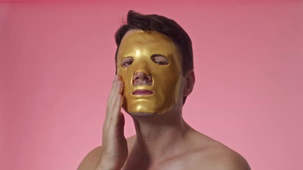 Testa di studio colpo di uomo dai capelli corti con occhi marroni che indossano una maschera di foglio idratante dorato sul viso isolato su rosa - Filmati, video