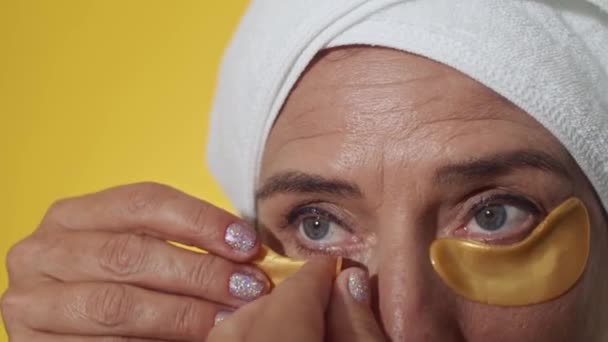Grande primo piano di donna matura con occhi blu e trucco leggero applicando patch idrogel sotto gli occhi isolati su sfondo giallo - Filmati, video