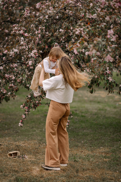 Όμορφη μητέρα και κόρη στο φόντο μιας ανθισμένης μηλιάς. Η μαμά μεγαλώνει την κορούλα της στην αγκαλιά της. Κομψά ρούχα σε ουδέτερα χρώματα. Η μαμά και η κόρη διασκεδάζουν. - Φωτογραφία, εικόνα
