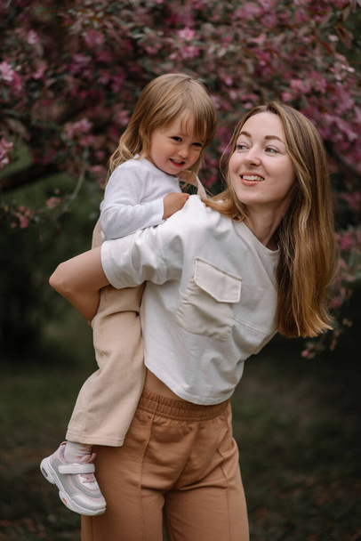 咲くリンゴの木の背景に対する美しい母と娘. 母は彼女の背中に小さな娘を乗せている. 中立色のスタイリッシュな服. ママと娘は楽しんでいる - 写真・画像