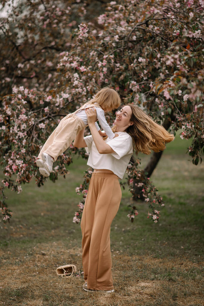 咲くリンゴの木の背景に対する美しい母と娘. ママは娘を腕の中で育てている. 中立色のスタイリッシュな服. ママと娘は楽しんでいる - 写真・画像