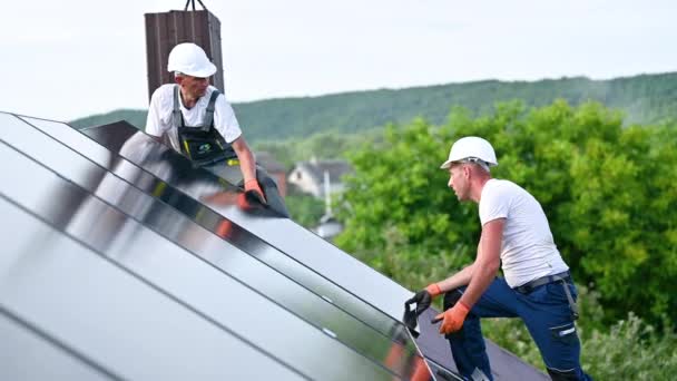 Los trabajadores que construyen el sistema de paneles solares en la azotea de la casa. Dos hombres instaladores en cascos instalando módulo solar fotovoltaico al aire libre. Concepto de generación de energía alternativa, verde y renovable. - Metraje, vídeo