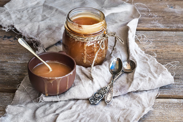 Salted caramel sauce in a rustic glass jar and brown ceramic cup on wooden desk - Fotó, kép