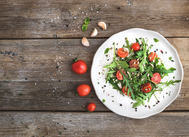 Salad with arugula, cherry tomatoes, sunflower seeds and herbs on white ceramic plate over rustic wood background, top view - Foto, afbeelding