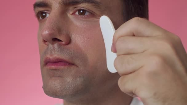 Close-up van mannelijke duizendjarige met bruine ogen doen anti-age massage met gua sha geïsoleerd op roze achtergrond - Video