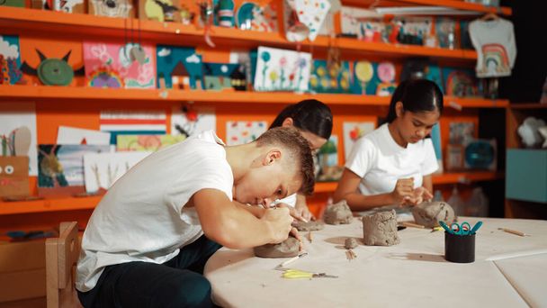 Adolescente de la escuela secundaria que usa una herramienta de tallado trabajando en arcilla en un taller de cerámica. Grupo de niños felices diversos trabajando o modelando taza de arcilla con la masa y el equipo de dispersión alrededor. Edificación. - Foto, imagen