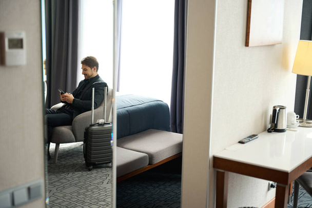 El hombre con un traje de viaje se sienta con un teléfono móvil en la silla, al lado de una maleta de viaje - Foto, imagen