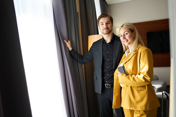 Casal viajante em pé junto à janela em um quarto de hotel, mulher em um terno de viagem amarelo - Foto, Imagem