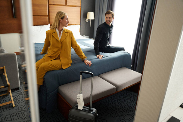 Reizend getrouwd stel ontspannen op bed in hotelkamer, vrouw in geel reispak - Foto, afbeelding