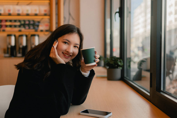 Retrato de menina coreana bonito com dois cafés em copos de papel no café perto da janela. Adolescente tendo cappuccino e olha para a câmera. - Foto, Imagem