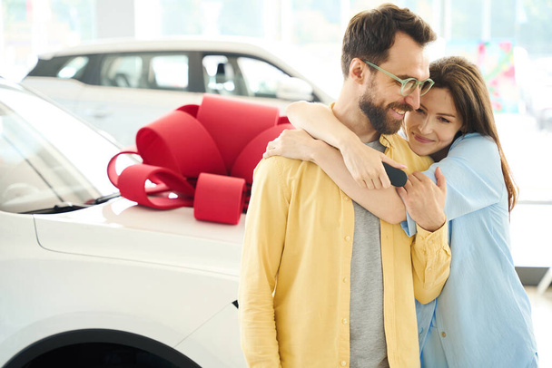 Šťastná žena objímající svého milovaného manžela, pár stojící v blízkosti auta s dárkovou mašlí - Fotografie, Obrázek