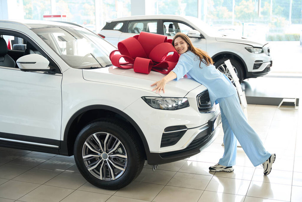 Mujer alegre abrazando el capó de un coche con un arco de regalo, ella está en un concesionario de automóviles - Foto, Imagen