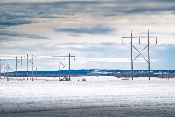Línea de grandes postes de energía de madera que sostienen líneas de transmisión a través de campos agrícolas cubiertos de nieve con vistas a colinas onduladas en el oeste de Canadá en Rocky View County Alberta. - Foto, imagen