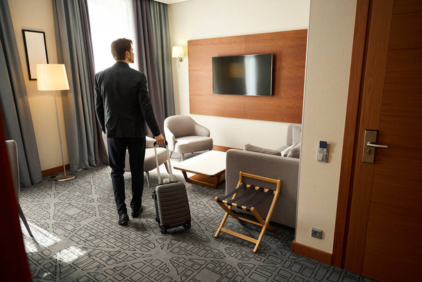Chico con una maleta de viaje se encuentra en una habitación de hotel, la habitación tiene un diseño minimalista moderno - Foto, imagen