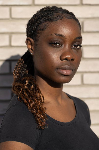 Dit boeiende beeld toont een close-up van een jonge Afrikaanse vrouw met een zelfverzekerde en directe blik. Het natuurlijke daglicht accentueert haar gelaatstrekken en de ingewikkelde vlechten in haar haar, die - Foto, afbeelding