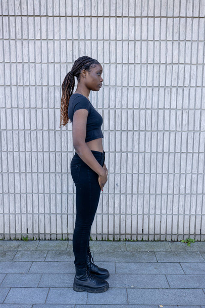 Esta fotografia apresenta um perfil lateral de uma jovem africana de pé contra um fundo de parede de tijolo uniforme. Seu olhar é direcionado para longe da câmera, o que adiciona um elemento de introspecção ou - Foto, Imagem