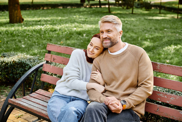 Ένα ερωτευμένο ζευγάρι με περιστασιακή ενδυμασία κάθεται μαζί σε ένα παγκάκι σε ένα ήσυχο περιβάλλον πάρκου. - Φωτογραφία, εικόνα
