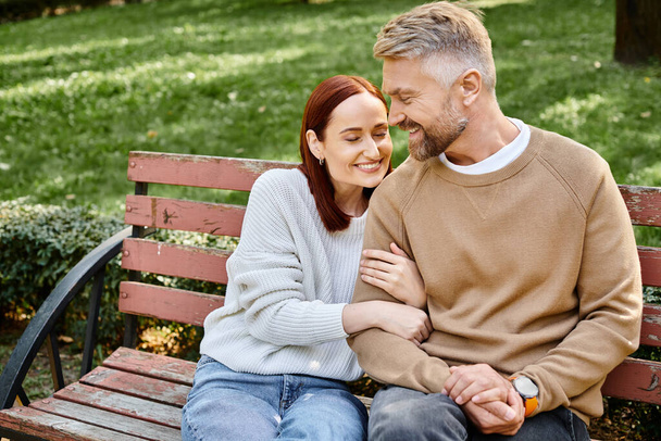 Een man en een vrouw in casual kleding zitten samen op een houten bank in een park. - Foto, afbeelding