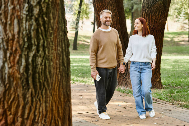 A man and woman in casual attire walk together in a peaceful park setting. - Fotoğraf, Görsel