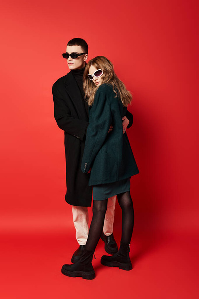 sophisticated young couple in stylish coats with sunglasses posing together on red backdrop - Zdjęcie, obraz