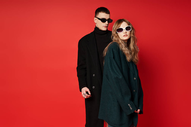 well dressed young couple in stylish coats with sunglasses posing together on red backdrop - Фото, зображення