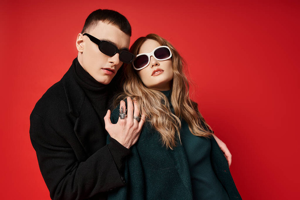 elegant young couple in stylish coats with trendy  sunglasses posing together on red backdrop - Foto, Bild