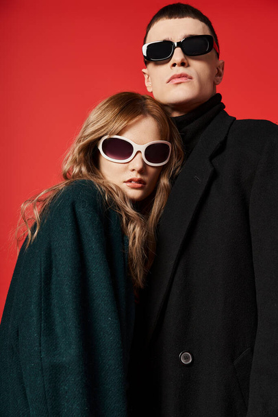 Şık ceketli, güneş gözlüklü genç bir çift kırmızı arka planda birlikte poz veriyorlar. - Fotoğraf, Görsel