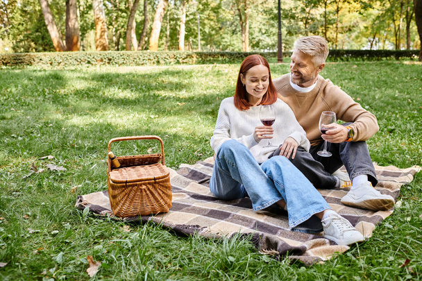 Um homem e uma mulher sentam-se em um cobertor na grama, desfrutando de um dia tranquilo no parque. - Foto, Imagem