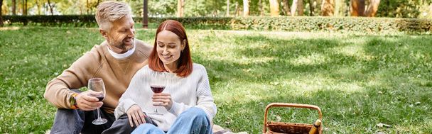 Uomo e donna adulti seduti su una coperta, che tengono bicchieri di vino in un parco. - Foto, immagini