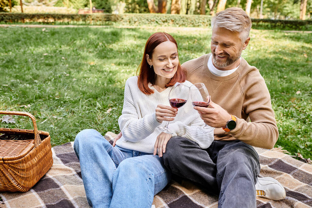 Un hombre y una mujer se sientan en una manta acogedora, sosteniendo copas de vino en un entorno romántico. - Foto, imagen