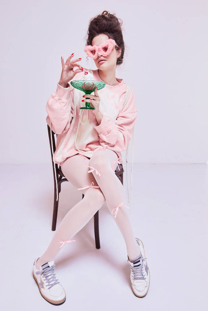 Young pretty woman sitting o chair in pink comfortable clothes and sunglasses, eating cake. Coziness at home. Life balance. Sweet tooth. Concept of beauty and fashion, vintage, boudoir style - 写真・画像