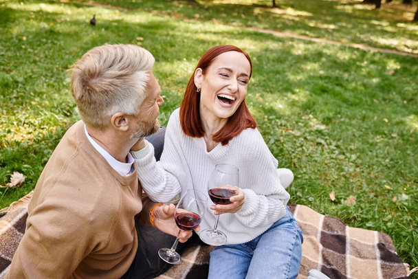 Un hombre y una mujer disfrutando de un picnic romántico mientras sostienen copas de vino. - Foto, imagen