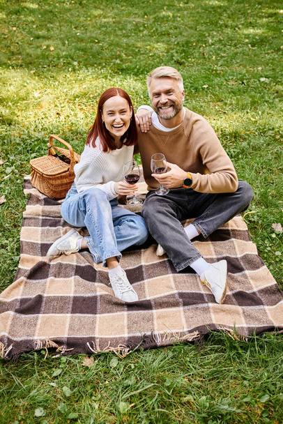 Ένα ζευγάρι κάθεται σε μια κουβέρτα, κρατώντας ποτήρια κρασιού σε ένα πάρκο. - Φωτογραφία, εικόνα